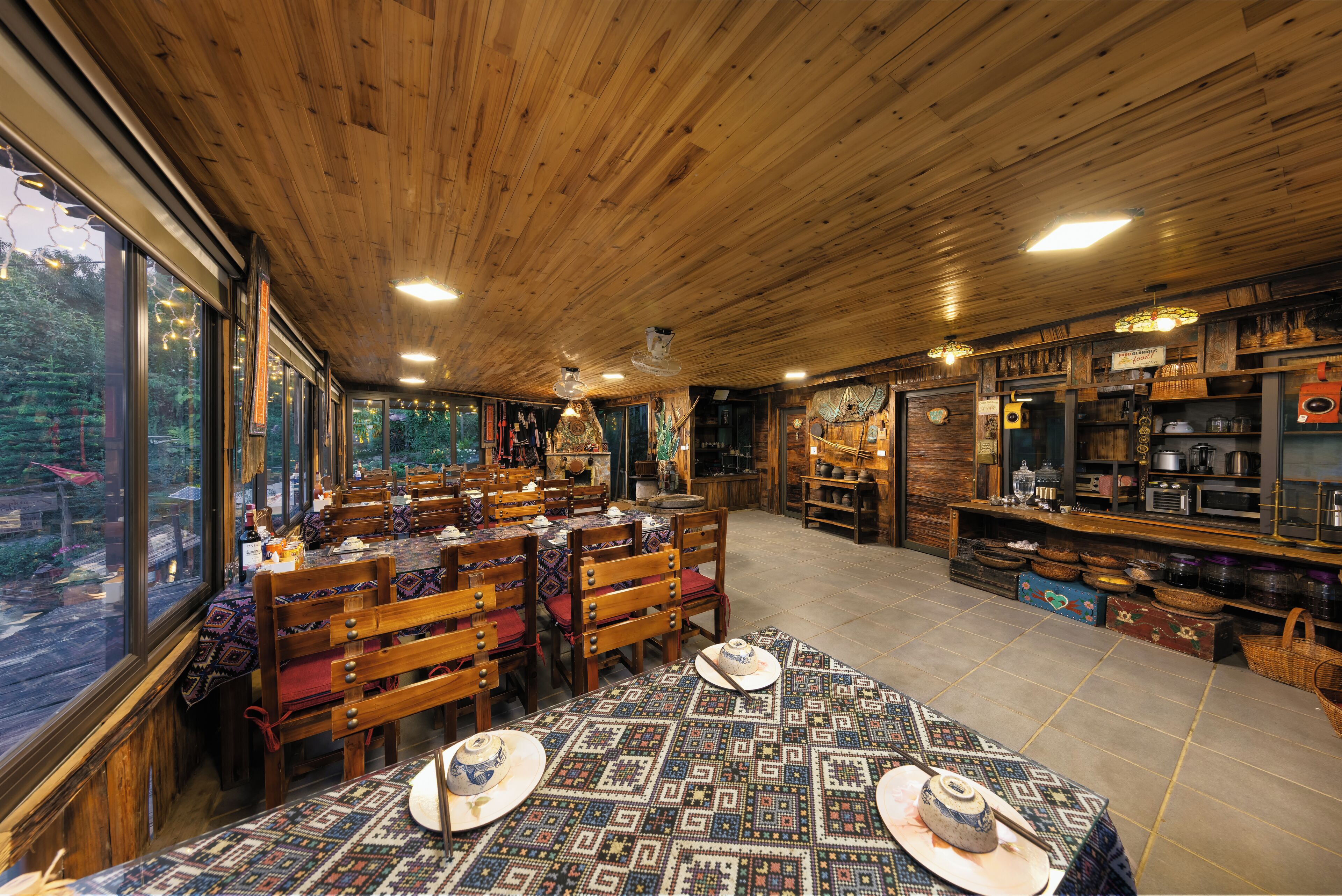 Interior rústico de madeira com mesas de jantar preparadas para hóspedes, grandes janelas com vista para o exterior e diversos utensílios de cozinha expostos em prateleiras.