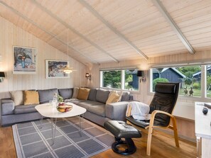 A modern living room with a sectional sofa, a round coffee table, a black leather chair, and a dining table. There are two windows providing a view of the outdoors.