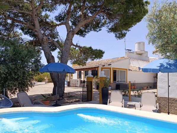 A pool area with lounge chairs and umbrellas, surrounded by trees and a building with a tiled roof.