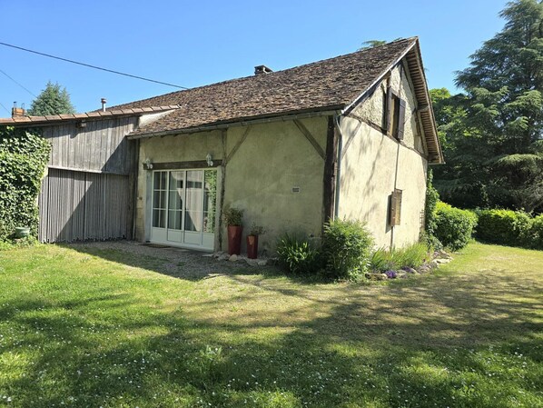 Una casa rústica de dos pisos, con techo inclinado, puerta principal con paneles de vidrio y un pequeño jardín con plantas en macetas.