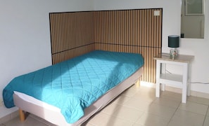A single bed with a blue quilt, a wooden slatted headboard, a white bedside table with a lamp, and a small shelf.