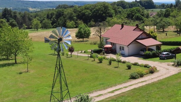 A rural house with a windmill, surrounded by greenery and a clear sky.