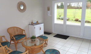 A room with wicker furniture, a glass table, a white cabinet, and a view of a green landscape through a door.