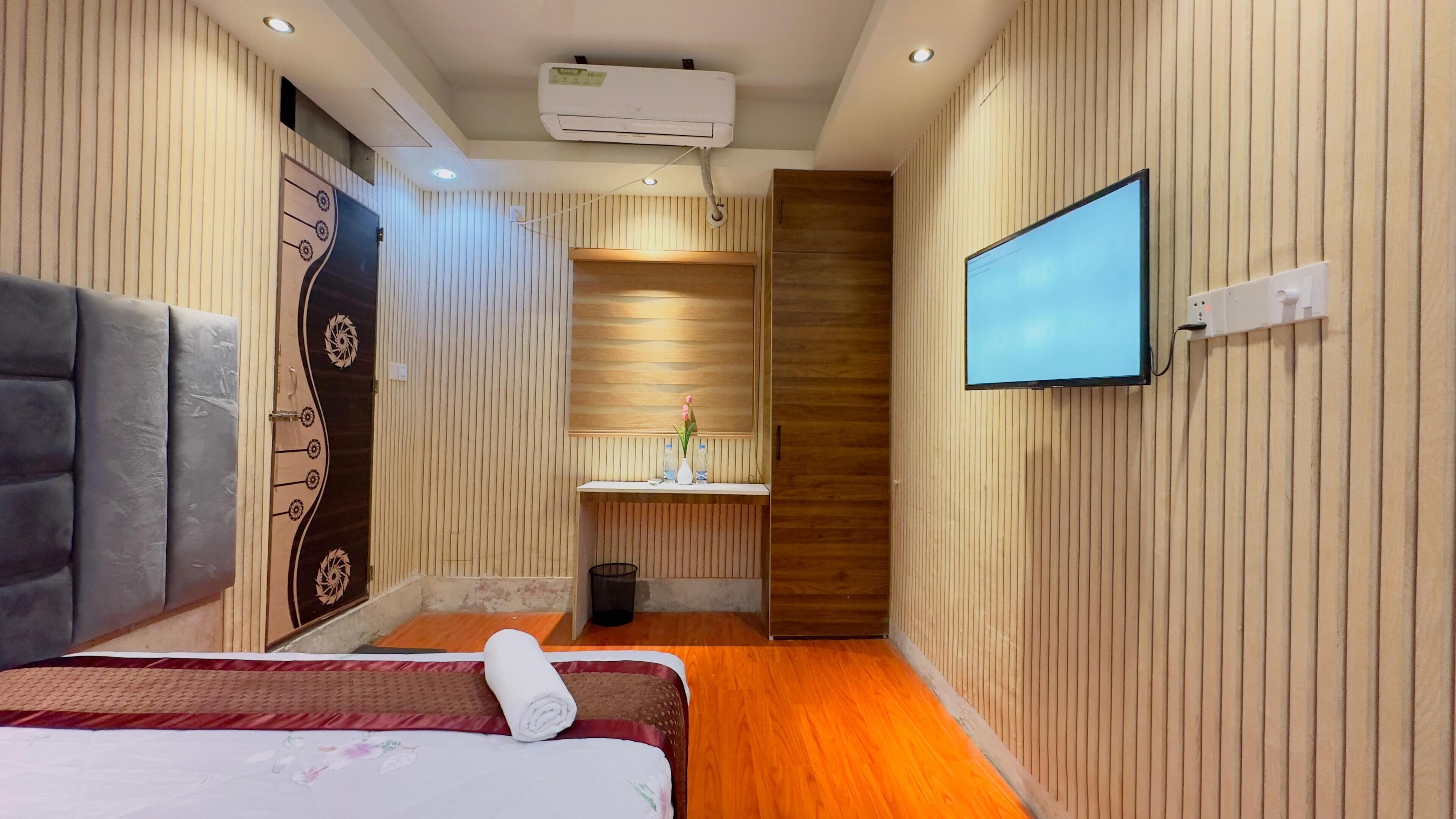 Ein Hotelzimmer mit einem großen Bett, einem an der Wand befestigten Fernseher, einem kleinen Badezimmer mit einem Waschbecken und einer Decken-Klimaanlage.