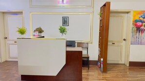 Un espace d’accueil avec un comptoir en bois, deux plantes en pot, un tableau encadré au mur et une peinture colorée sur le mur adjacent.