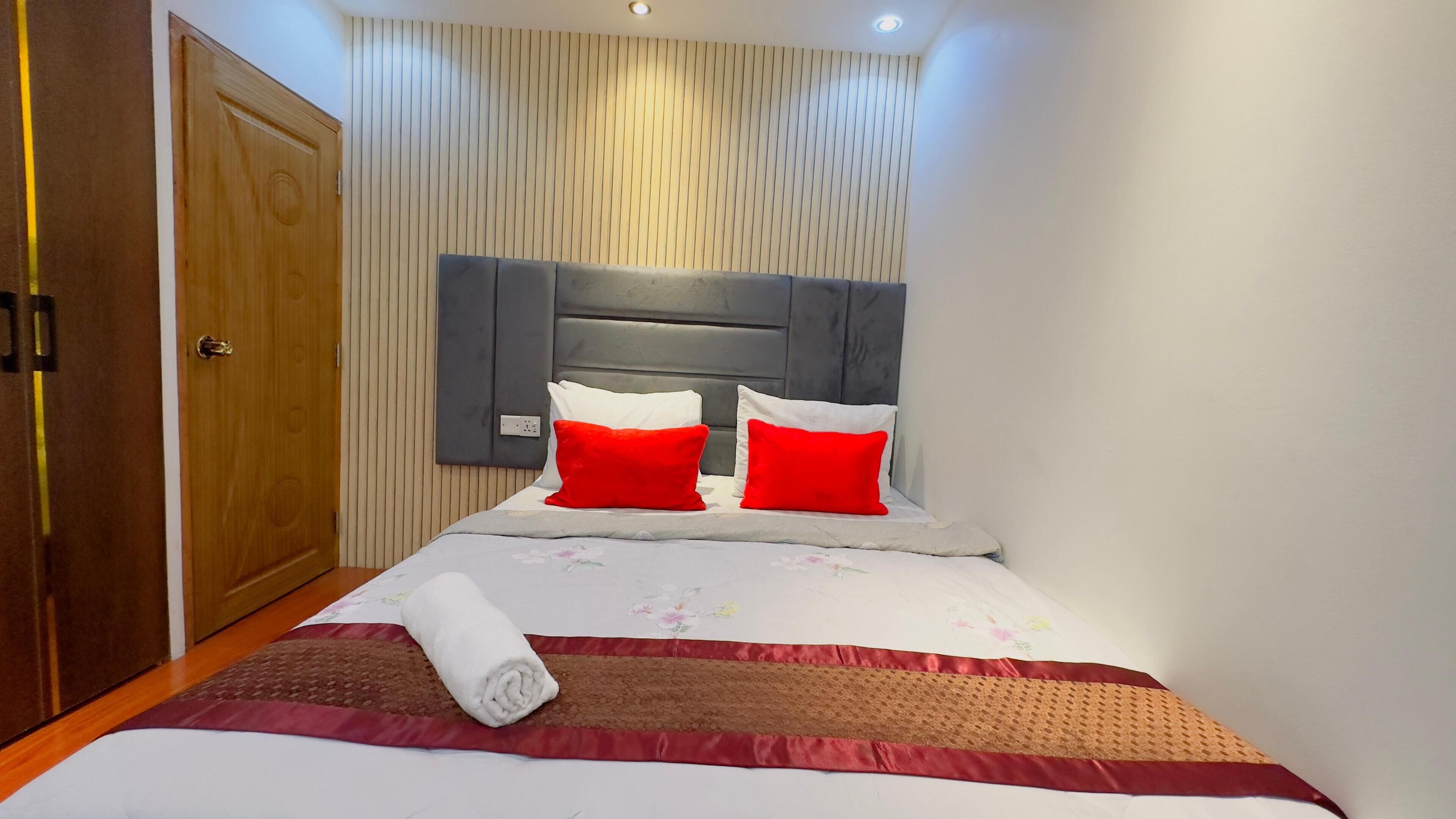 A neatly made bed with a red and white pillow set, a rolled towel, and a floral-patterned bedspread.