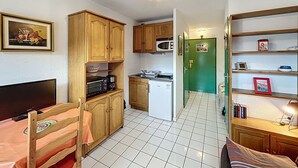 A compact kitchen with wooden cabinets, a microwave, a refrigerator, and a dining area with a chair and table. - Bright studio with balcony, pets allowed (Lamalou-les-Bains)