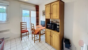 A room with a dining table, chairs, a wooden cabinet with kitchen appliances, a window with curtains, and a ceiling light fixture. - Bright studio with balcony, pets allowed (Lamalou-les-Bains)