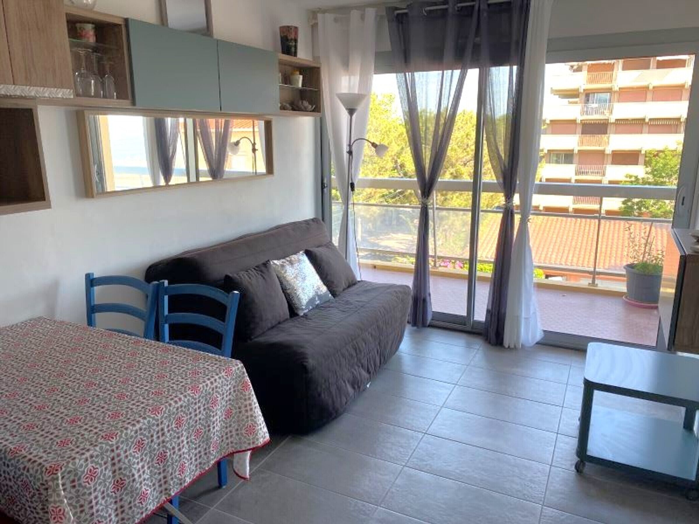 A furnished room with a sofa, a table, and a chair, a balcony with a view of buildings, and a window with curtains.