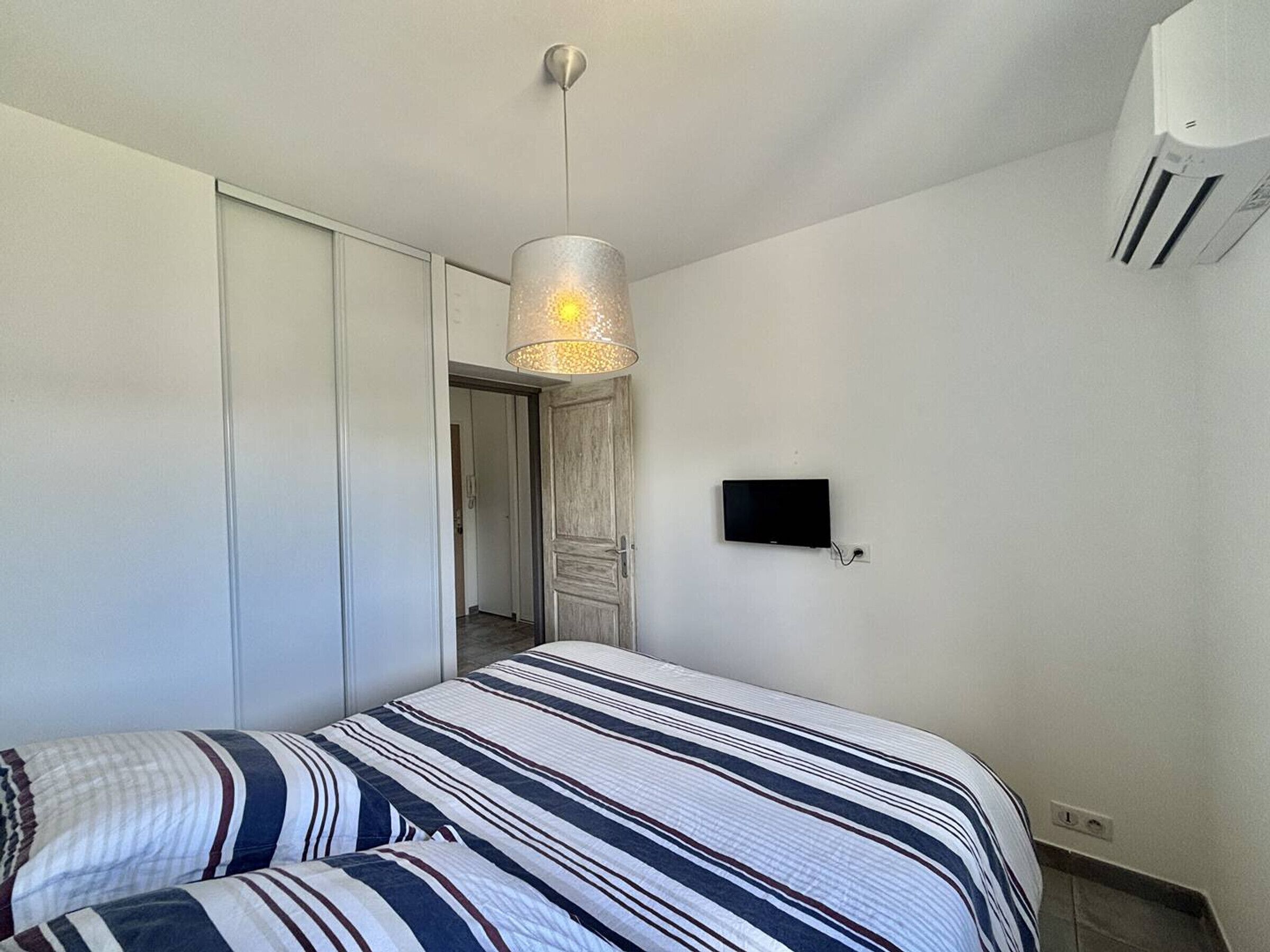 A bedroom with a bed, a hanging light fixture, a TV mounted on the wall, and a wardrobe.