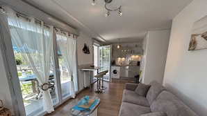 Una sala moderna con un sofá gris, una mesa de centro y una zona de comedor con una mesa y sillas. Se ve una cocina al fondo con lavadora y una mesa de comedor.