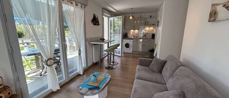 A modern living room with a grey sofa, a coffee table, and a dining area with a table and chairs. There is a kitchen in the background with a washing machine and a dining table.