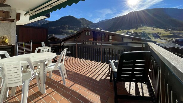 Una terraza con mesa y sillas, vistas a montañas y un cielo soleado.