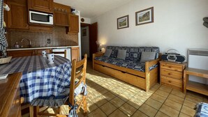 Cocina compacta con armarios de madera, microondas y una mesa de comedor con mantel a cuadros. Hay un sofá con cojines estampados y una radio sobre una mesita de madera.