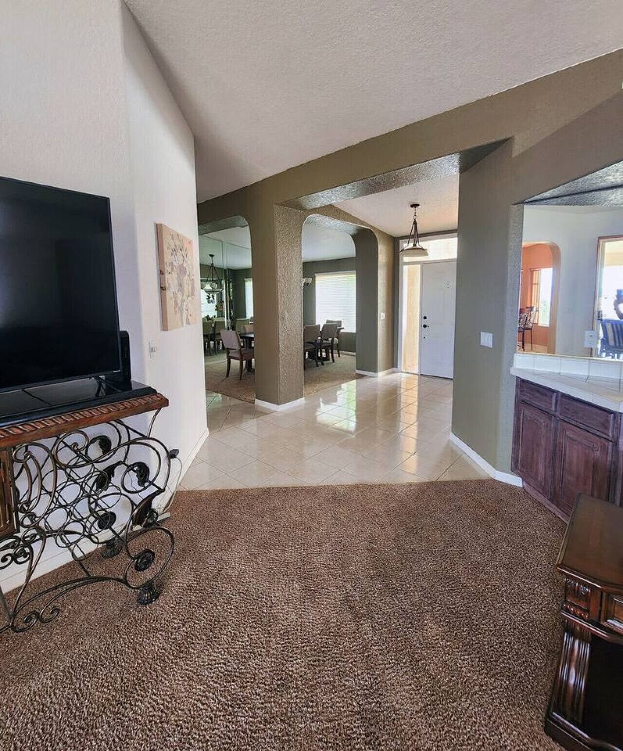 A spacious living room with a flat-screen TV on a stand, a dining area with chairs and a table, and a kitchen area with cabinets and a countertop.