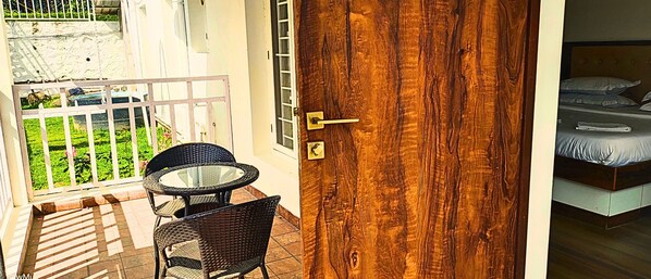 A balcony with wicker chairs and a glass table, a wooden door with a brass handle, and a bed with pillows. - The Summit Homestay (Kodaikanal)