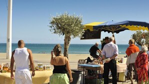 Ein Sitzbereich im Freien direkt am Strand mit Tischen und Stühlen, einem großen Sonnenschirm und Blick auf das Meer.
