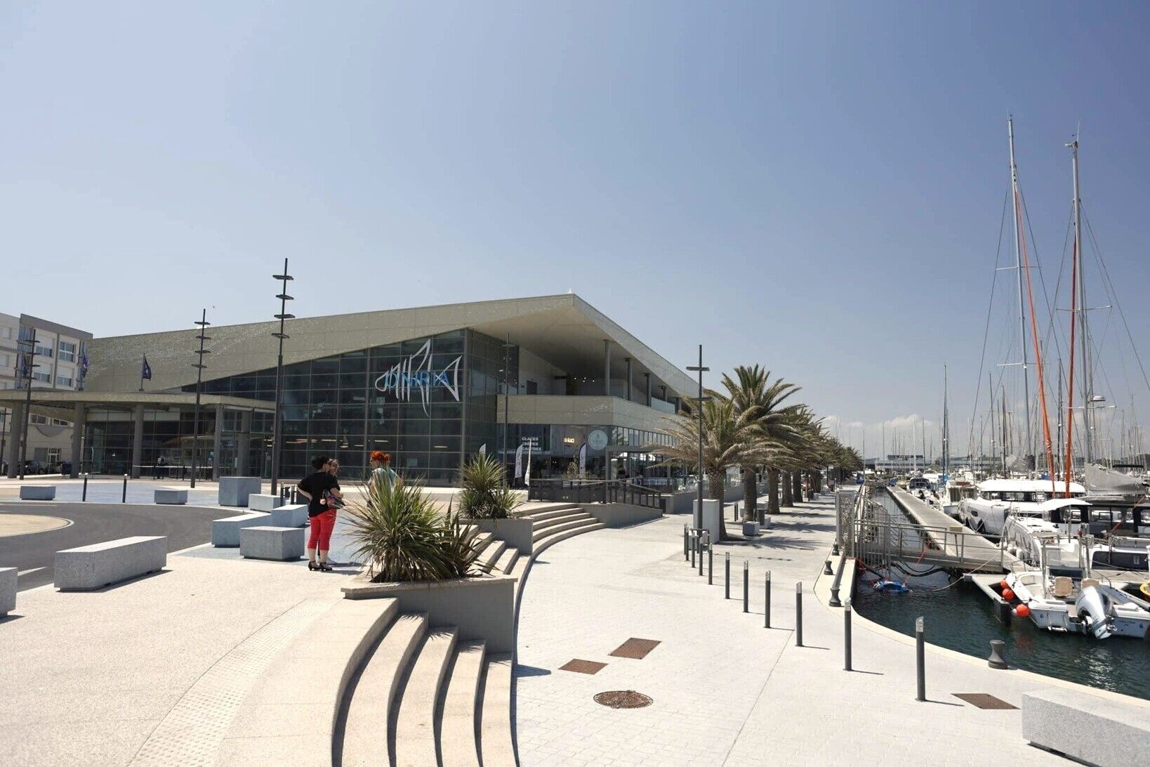A waterfront promenade with a modern building, palm trees, and a marina with boats.