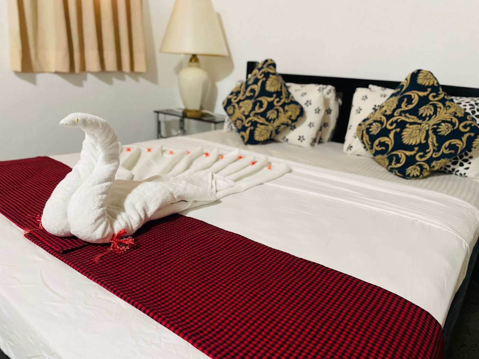 Una cama bien hecha con una colcha a cuadros rojos y blancos, almohadas blancas con diseños dorados y una toalla blanca con forma de cisne.