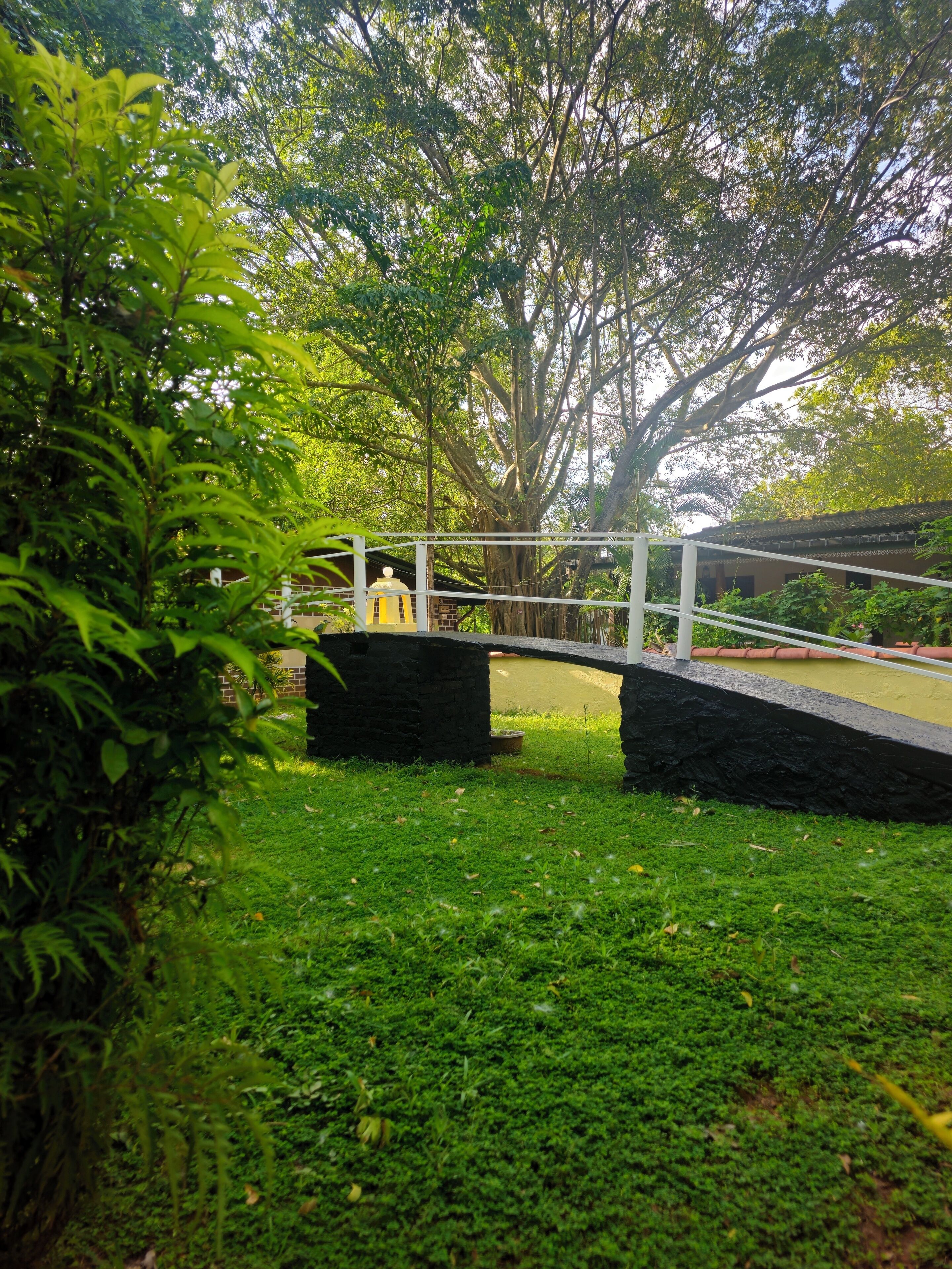 A stone bridge over a grassy area with a large tree and a building in the background.