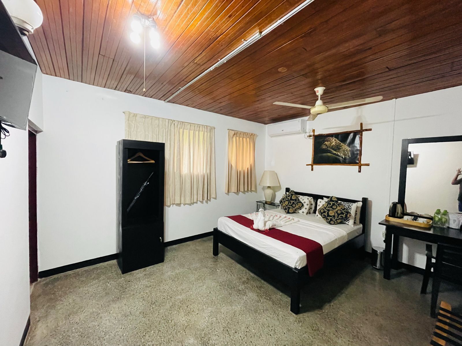 A bedroom with a bed, a wardrobe, a ceiling fan, a painting, and a mirror.