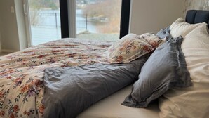 A bedroom with a bed, floral bedding, and a view of a lake and mountains through a large window. - Villa by the sea with access to sea shed and decking (Vestland)