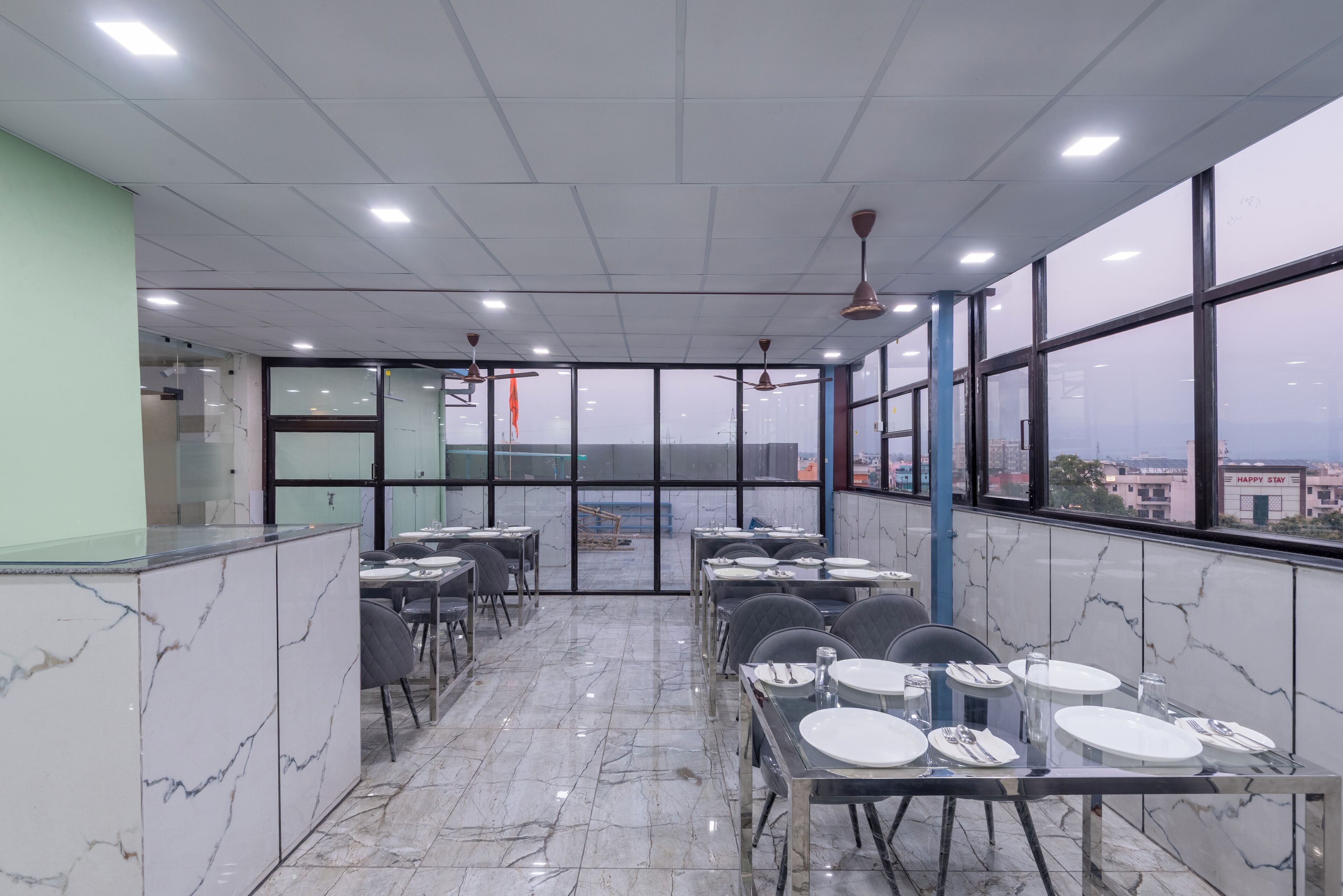 Un restaurant doté d’un sol en marbre, de tables et de chaises en verre, de grandes fenêtres offrant une vue dégagée et de ventilateurs de plafond.