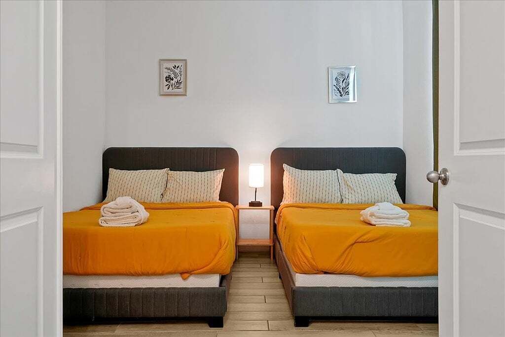A hotel room with two beds, each with an orange bedspread and white pillows. There is a nightstand between the beds with a lamp on it. The room has white walls and a wooden floor.