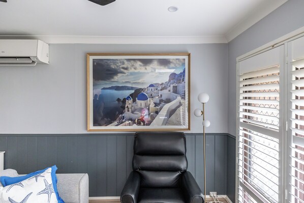 A room with a framed painting of a coastal town, a black leather chair, a white air conditioner, and a window with blinds.