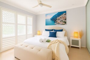 Quarto com cama, duas luminárias de cabeceira, ventoinha no teto, janela com persianas e uma pintura de uma cidade costeira.
