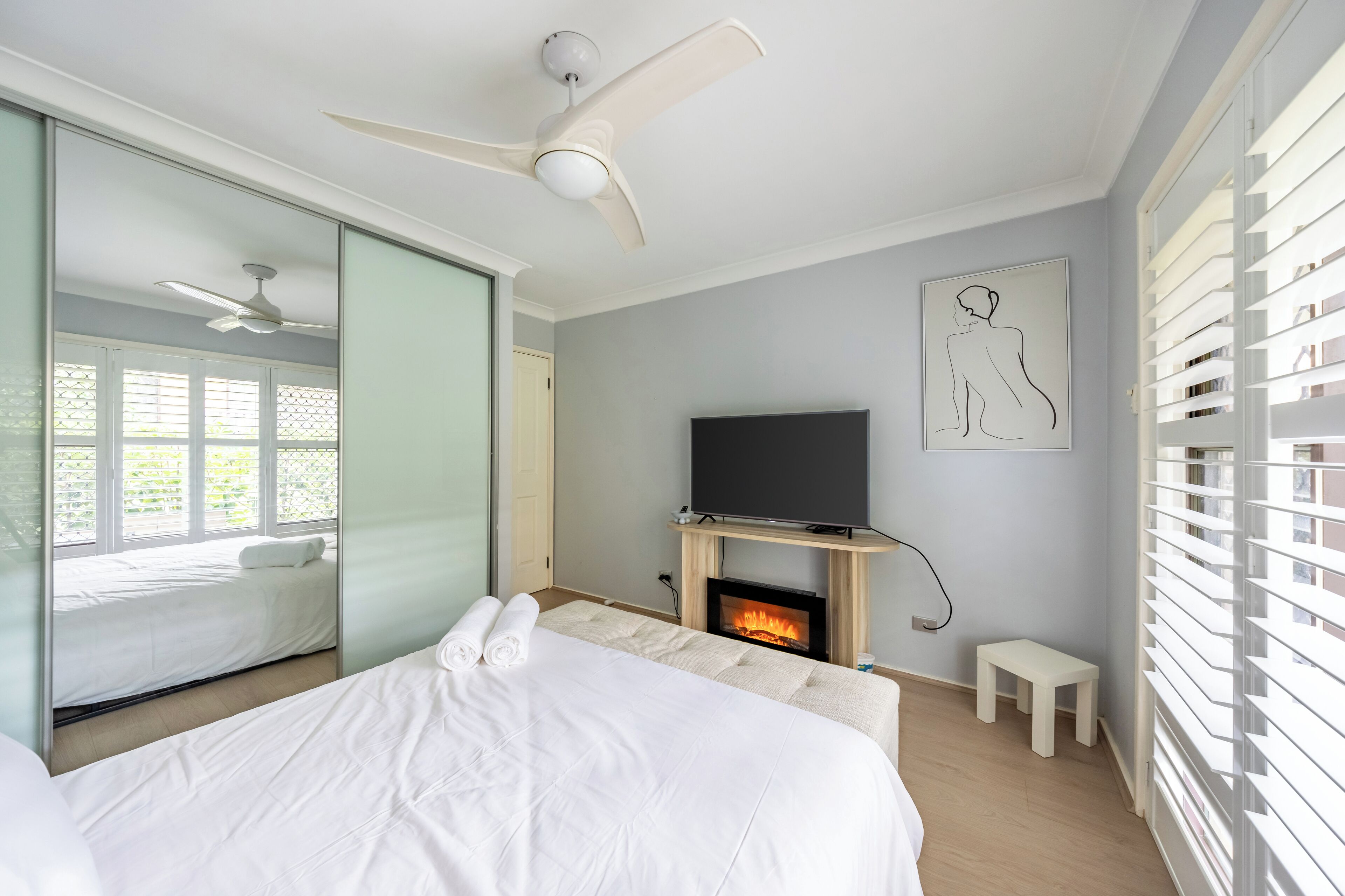 A bedroom with a bed, a television, a fireplace, a ceiling fan, and a window with blinds.