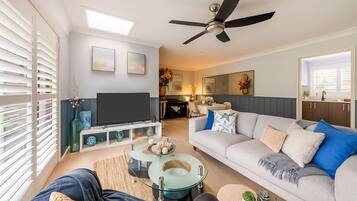 A modern living room with a flat-screen TV, a glass coffee table, and a grey sofa with blue cushions.