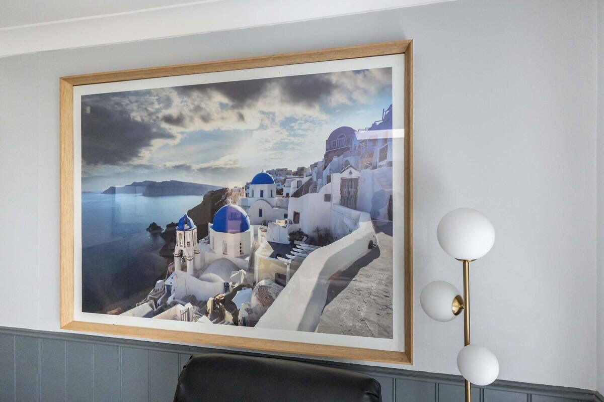 Un quadro incorniciato raffigura una cittadina costiera con edifici bianchi e cupole blu, una lampada da terra moderna e una sedia.