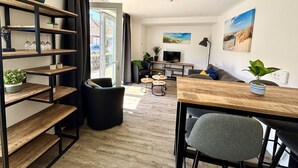 Un salon moderne avec une table à manger en bois, des canapés noirs et un téléviseur. On y trouve des étagères avec des plantes et une grande fenêtre avec des rideaux.