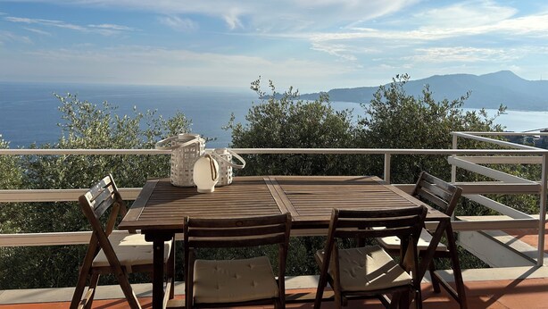 Ein Holztisch mit Stühlen auf einer Terrasse mit Blick auf das Meer und ferne Berge.