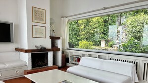 Ein gemütliches Wohnzimmer mit Kamin, einer Couch, einem Couchtisch mit Zeitschriften und einem großen Fenster mit Blick auf Grünfläche.