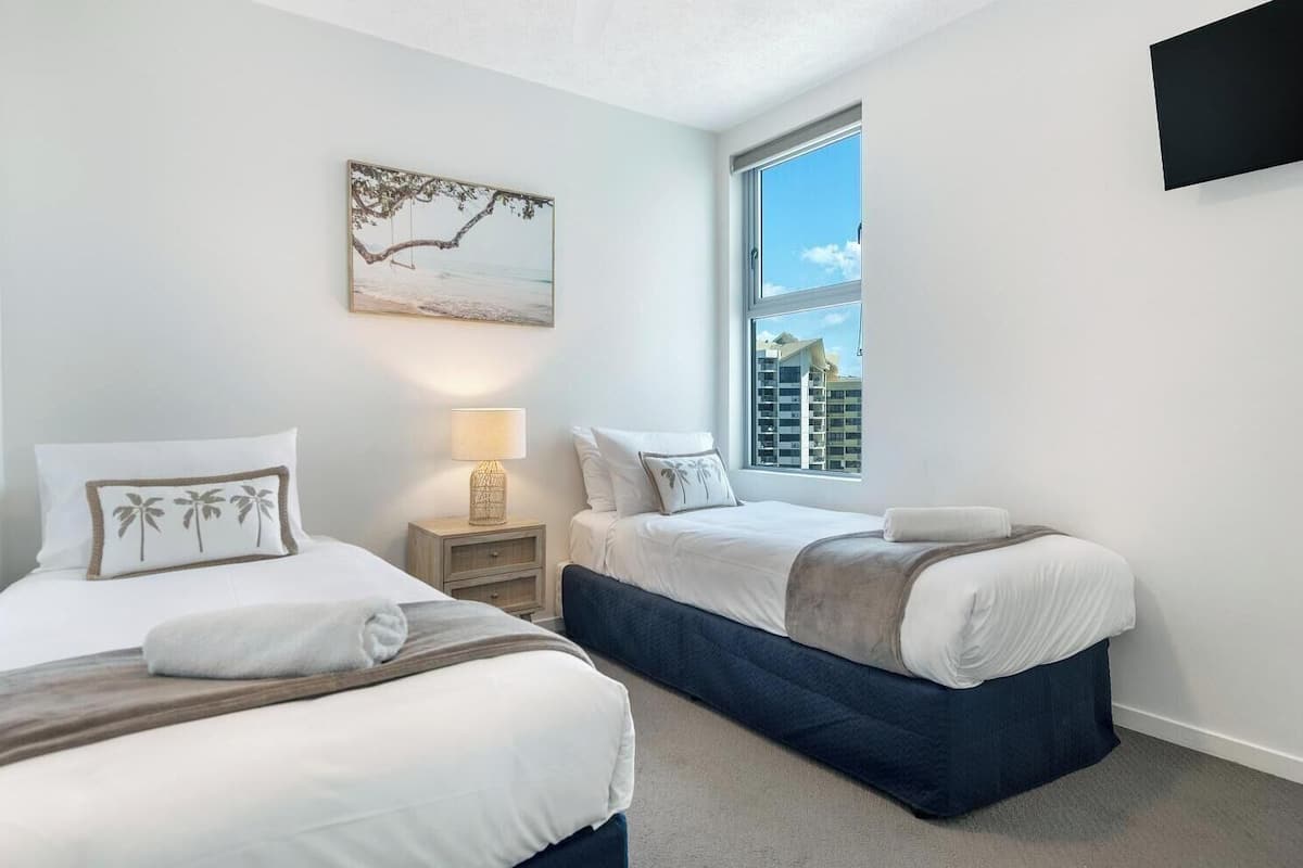 A hotel room with two beds, a nightstand with a lamp, a painting of a beach scene, and a window with a view of buildings.