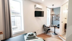 Ein modernes Hotelzimmer mit Bett, Schreibtisch und Stuhl. Es gibt ein Fenster mit Vorhängen, einen an der Wand montierten Fernseher und eine Deckenleuchte.