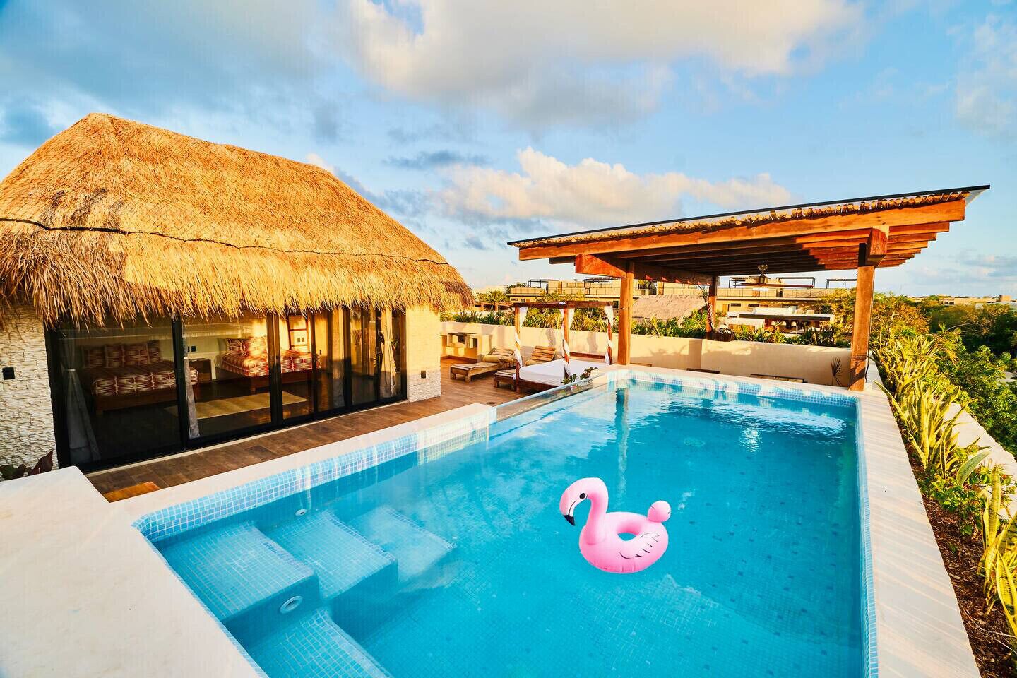 A swimming pool with a thatched-roof structure and a wooden pergola, surrounded by lush greenery.