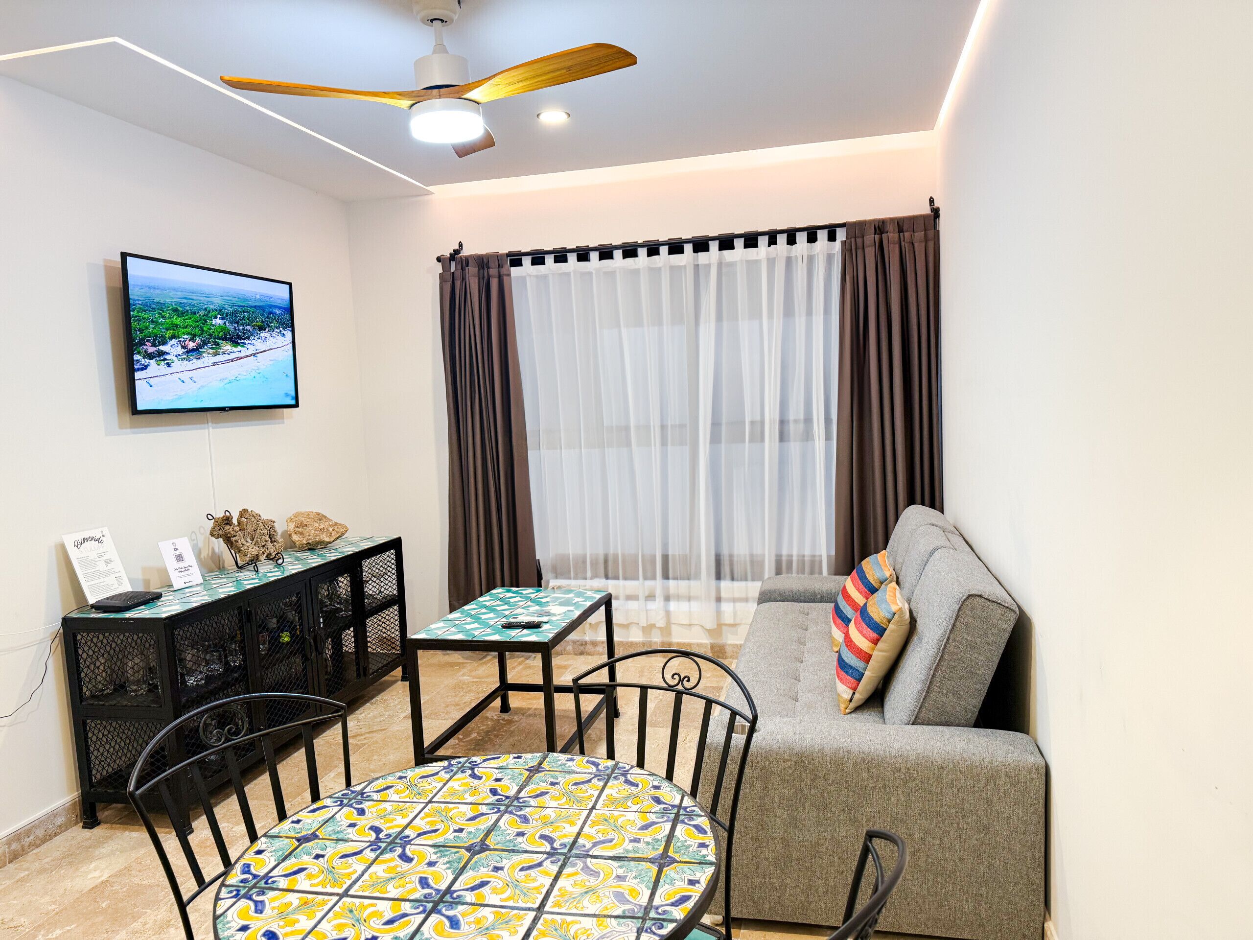 Uma sala de estar moderna com um sofá cinzento, uma mesa redonda com toalha de mesa estampada, um ventilador de teto e uma TV na parede.