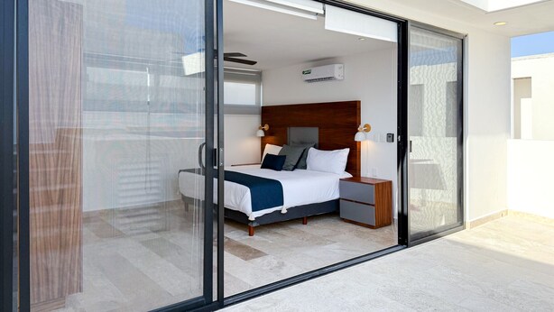 Een moderne slaapkamer met een groot schuifglasraam dat leidt naar een balkon, een houten hoofdbord, een nachtkastje en een wandairconditioner.