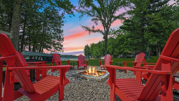 A fire pit surrounded by red Adirondack chairs in a park setting with trees and a lake in the background. - Sand Bar Resort – Updated 5BR with Lake Views,  Big Yard, Firepit, Pontoon rental,  Gourmet Kitchen & More! (Suring)