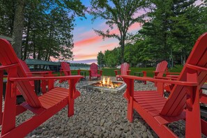 A fire pit surrounded by red Adirondack chairs in a park setting with trees and a lake in the background. - Sand Bar Resort – Updated 5BR with Lake Views,  Big Yard, Firepit, Pontoon rental,  Gourmet Kitchen & More! (Suring)