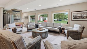 A spacious living area with a large sofa set, a round coffee table, and a view of a green landscape through the windows. - Sand Bar Resort – Updated 5BR with Lake Views,  Big Yard, Firepit, Pontoon rental,  Gourmet Kitchen & More! (Suring)