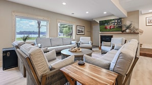 A spacious living room with a large sofa set, a coffee table, and a fireplace. There are large windows providing a view of the outdoors. - Sand Bar Resort – Updated 5BR with Lake Views,  Big Yard, Firepit, Pontoon rental,  Gourmet Kitchen & More! (Suring)