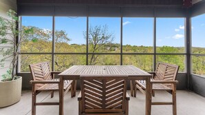 A covered patio with a wooden table and chairs, large windows offering a view of trees and hills.
