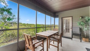 A covered patio with a wooden table and chairs, large windows offering a view of the surrounding landscape.