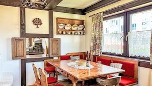 Un comedor con una mesa de madera, bancos tapizados en rojo y una ventana con cortinas de encaje.