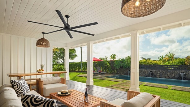A covered outdoor seating area with a ceiling fan, a wooden table with a bottle and glasses, and a view of a swimming pool and garden.