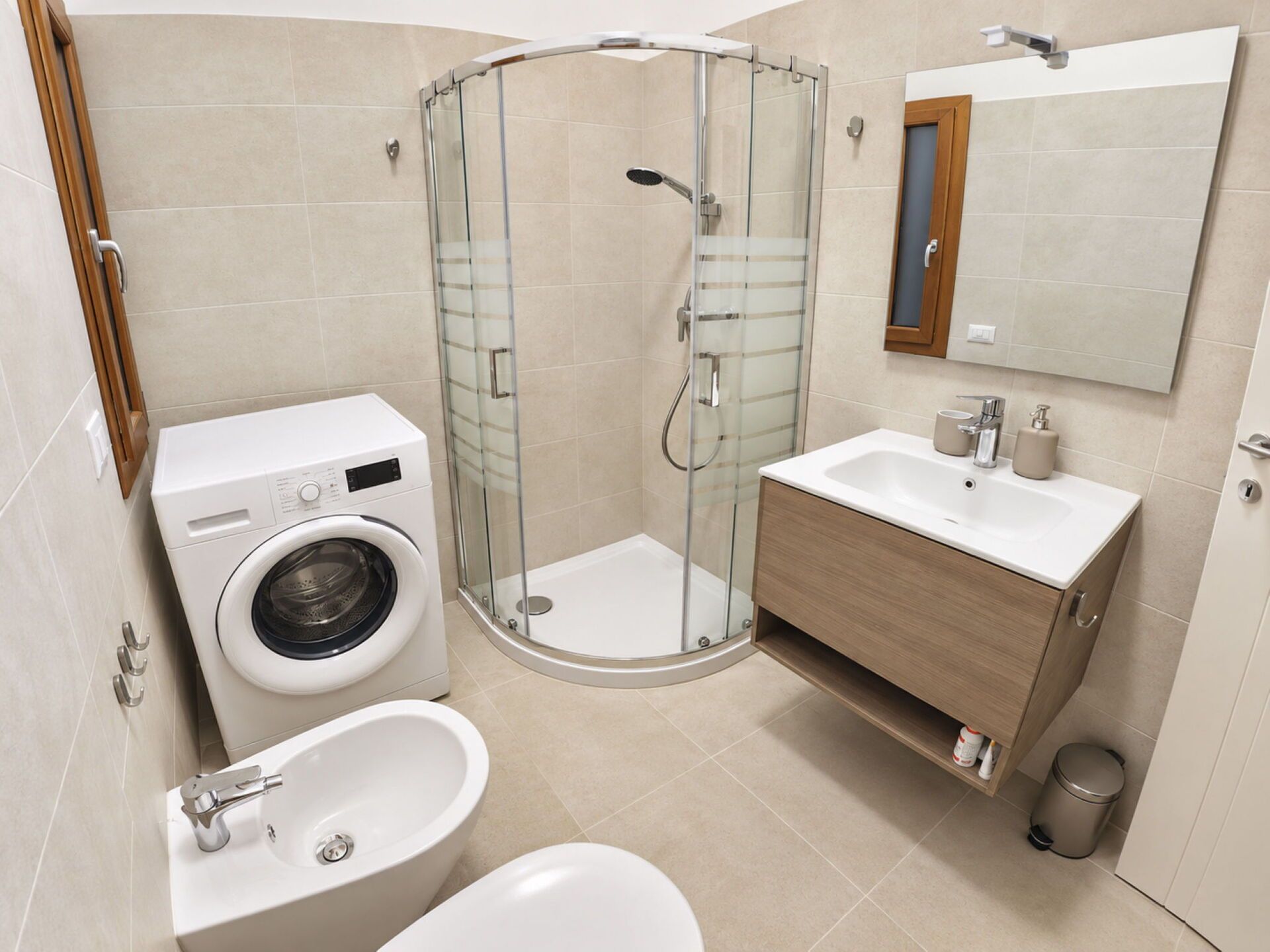 A modern bathroom with a glass shower enclosure, a built-in washing machine, a sink, and a vanity with a mirror.